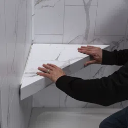 Hands installing a white marble-patterned triangular shelf in a tiled corner bathroom wall above a bathtub.
