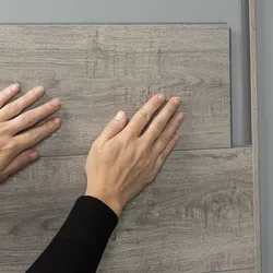 Hands installing gray wood-look laminate flooring planks over a gray subfloor surface indoors