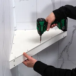 Person installing a white corner shelf using a green cordless drill against a marble tiled wall.