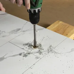 Handheld electric drill making a hole in white marble tile with dust and debris scattered around.