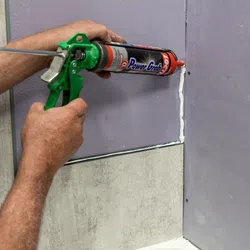 Hands applying white sealant with green caulking gun on purple wall panels and concrete surface corner.