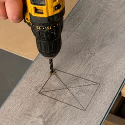 Person using a yellow and black drill to make a hole in a marked rectangular shape on a wooden plank.
