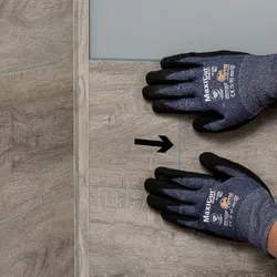 Hands wearing MaxiCut safety gloves placing wood-look floor tiles with alignment arrow on surface.