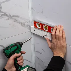 Person using a green cordless drill to screw a white wooden board to a tiled wall, checking level with a red spirit level.