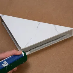 Hand applying adhesive with a caulking gun to the underside of a triangular marble-patterned tile on a brown surface.