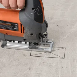 Close-up of a hand holding an orange electric jigsaw cutting a rectangular hole in a concrete surface.