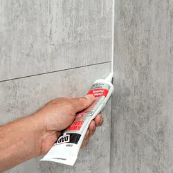 Hand applying white sealant to bathroom corner between bathtub and tiled wall for waterproofing.
