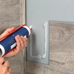 Hands applying white adhesive with a caulking gun on a wall next to wood-patterned tiles for installation.