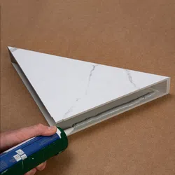Hand applying adhesive with caulking gun to secure white marble-patterned triangular panel on brown surface.