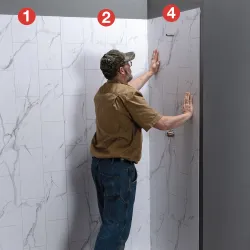 Man installing large white marble-effect wall panels in a corner bathroom space with gray walls.