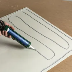 Hand holding a caulking gun applying grey adhesive in wavy lines on a white board set on brown surface.