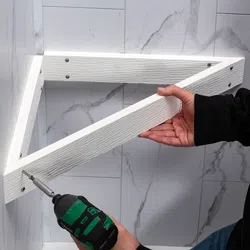 Person using a cordless drill to install a white triangular wooden shelf on a marble tiled wall.
