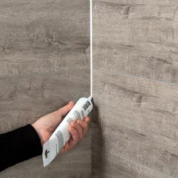 Person applying white sealant with caulking gun to the corner of wood-textured tiled wall.