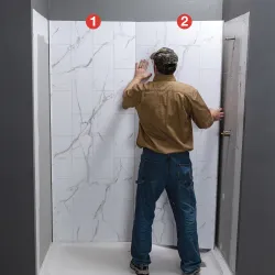 Man installing marble-patterned wall panels with numbered steps in a shower stall renovation.