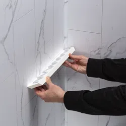 Hands installing a white corner tile shelf on a marble-patterned tiled wall in a bathroom or kitchen corner.