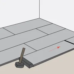 Installing gray wooden floor planks with a rubber mallet and tapping block in a staggered pattern in a room corner.