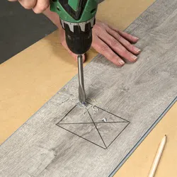 Person drilling a hole into a marked wooden plank using a green electric drill on a workbench.