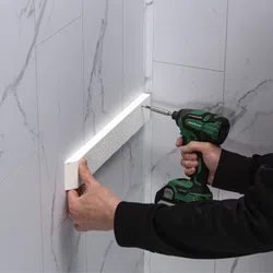 Person using a cordless drill to install a white textured trim piece on a marble-patterned tiled wall.