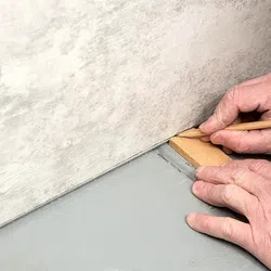 Hand marking a wooden floor trim piece with a pencil against a wall for precise fitting during installation.
