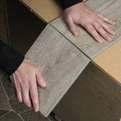 Person installing gray wood-look vinyl flooring planks on a wooden surface, hands aligning edges precisely.
