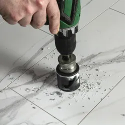 Hand using green electric drill with hole saw bit to drill through marble tile creating debris on surface.