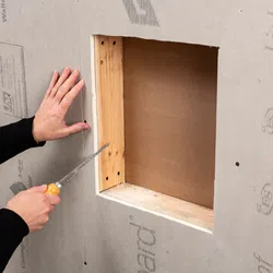 Person cutting drywall around a wooden window frame with a handsaw for installation or repair.