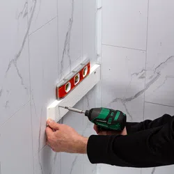 Person using a green cordless drill to screw a white textured panel on a wall with a red spirit level on top.