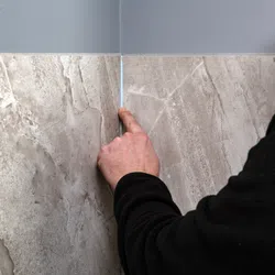 Person pointing at the thin gap between two beige marble wall tiles under a gray painted corner wall.