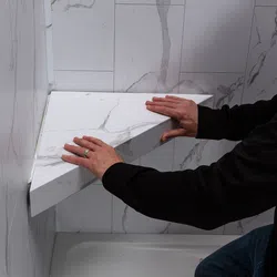 Person installing a triangular white marble corner shelf with gray veins in a tiled bathroom shower area.