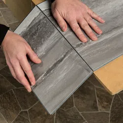 Person's hands installing gray wood-look vinyl flooring panels on a floor with patterned stone tiles.