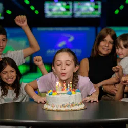 Girl blowing out candles on birthday cake surrounded by cheering family at a bowling alley party.