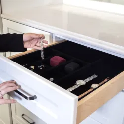 Woman organizing watches and jewelry in a white wooden drawer with black velvet organizer inside a modern kitchen.