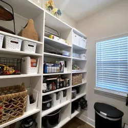 Organized pantry shelves with baskets, food items, appliances, and storage containers beside a window with blinds.