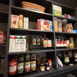 Organized pantry shelves with boxed pasta, broth cartons, canned goods, cereals, condiments, and jars in dark wooden shelving.