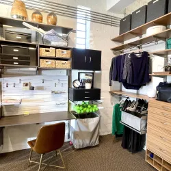 Organized walk-in closet with shelves, baskets, hanging clothes, shoes, bags, and a small workspace with chair and drawers.