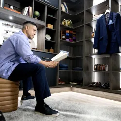 Modular storage systems vs custom solutions - Man sitting on a ottoman reading a book inside a modern walk-in closet with clothes and shoes.