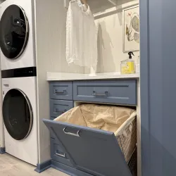 Stacked washer and dryer next to blue cabinet with pull-out laundry hamper and white hanging blouse in bright laundry room