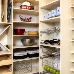 Organized pantry shelves with storage bins, spices, canned goods, metal bowls, and apples in wire baskets.
