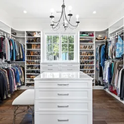 Bright spacious walk-in closet with white cabinetry, marble island, chandelier, and organized clothing and shoe racks.