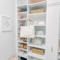 Organized white closet shelves with shoes, bags, wicker baskets, folded clothes, and decorative items in soft lighting