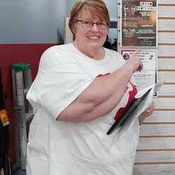 Smiling woman in white shirt pointing at a grand reopening sign inside a store with rifles on the wall.