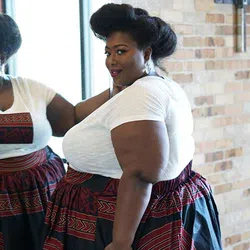 Confident plus-size woman in a white top and patterned skirt poses by mirror against brick wall.