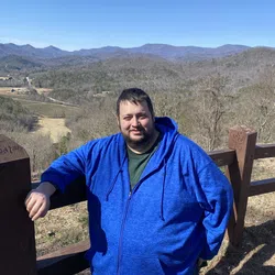 Man in blue hoodie standing by a wooden fence overlooking a valley with mountains under a clear blue sky.