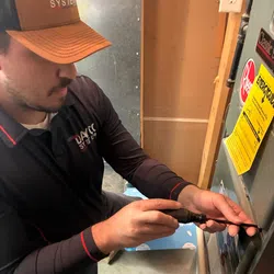 Technician wearing a brown cap and black shirt repairing an HVAC unit with a screwdriver indoors.