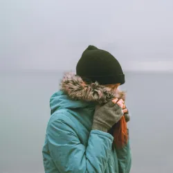 Person in a teal parka and black beanie covering face with gloved hands near a foggy ocean background.