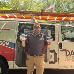 Dayco Systems HVAC technician holding parts in front of company van with ladder and American flag.