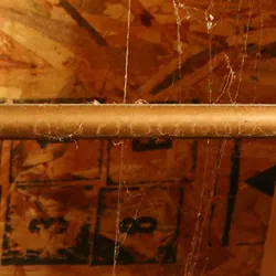 Close-up of an attic ceiling with wooden beams, a copper pipe, spider webs, and stamped numbers on plywood.