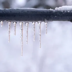 Clear icicles hanging from a snow-covered metal pipe in a cold winter setting with blurred background.