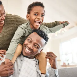 Happy African American family enjoying playful moment at home with child on father's shoulders and mother smiling.