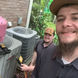 Two smiling HVAC technicians working outdoors on air conditioning units beside a brick house surrounded by greenery